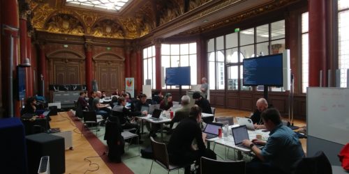 People clustered around 3 tables working on their computers in a gorgeous 19th century Parisian ballroom.