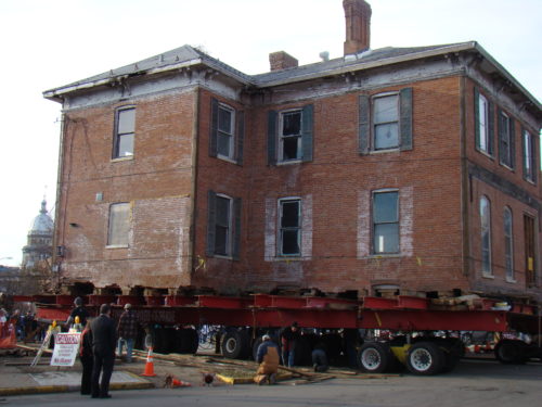 A two-story, 19th-century building, loaded on 64 wheels, is moved down a street by a dozen workers in hardhats